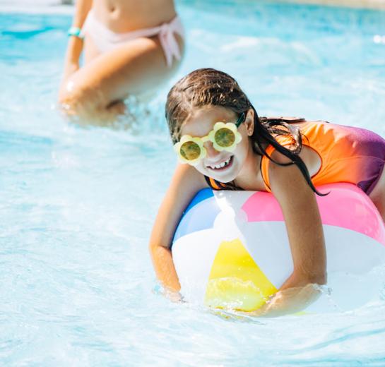 Bambina in piscina con occhiali e palla colorata.